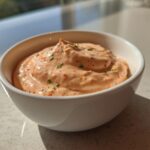A small white bowl filled with creamy, orange-hued Copycat Classic Burger Spread Sauce, garnished with herbs.