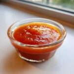 Close-up of vibrant orange Copycat Sweet-Tangy Polynesian Dipping Sauce in a small clear glass ramekin.
