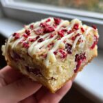 A hand holding a square of delicious cranberry bliss bars topped with white chocolate drizzle and dried cranberries.