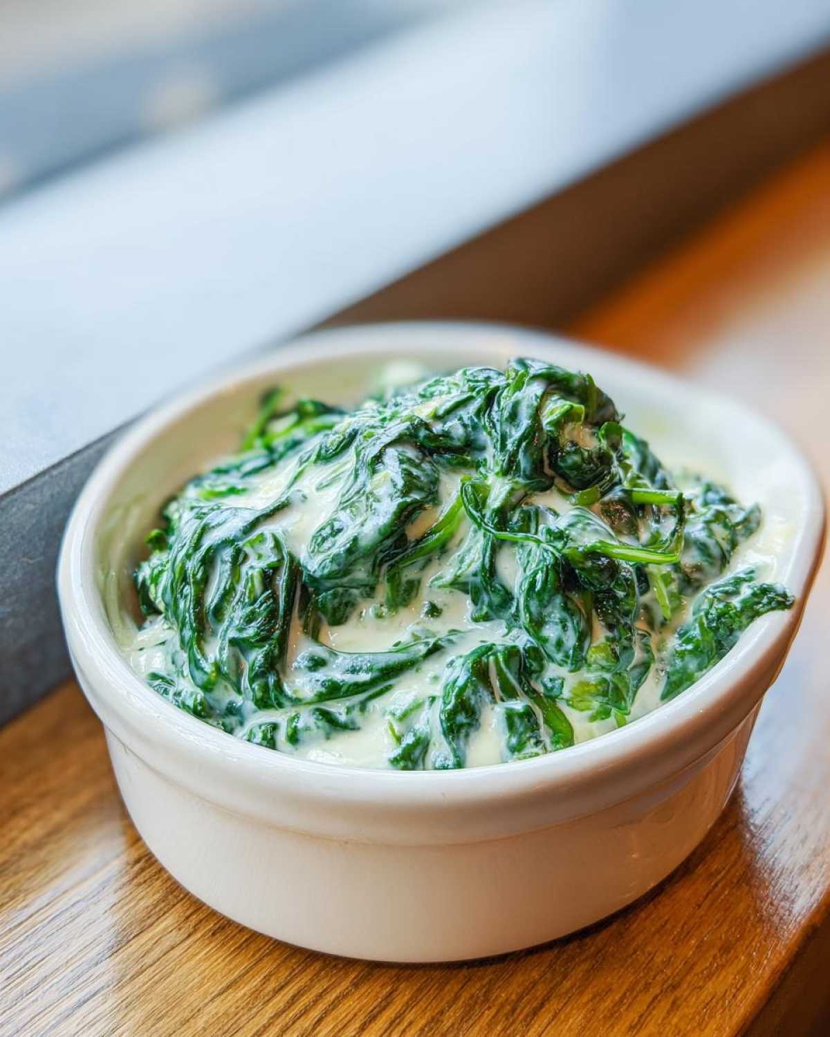 A close-up of rich, vibrant green creamed spinach served in a small white bowl on a wooden surface.