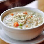 Close-up of a steaming white bowl filled with creamy chicken and rice soup, featuring shredded chicken, carrots, and peas.