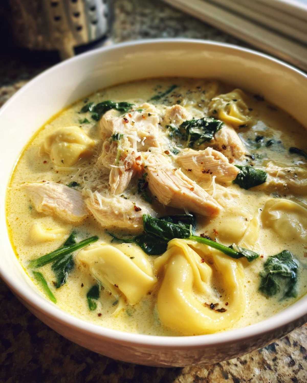 Close-up of a white bowl filled with creamy chicken tortellini soup, featuring chunks of chicken, spinach, and cheese garnish.