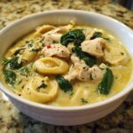 A close-up shot of a bowl of creamy chicken tortellini soup, featuring chunks of chicken, spinach, and pasta.