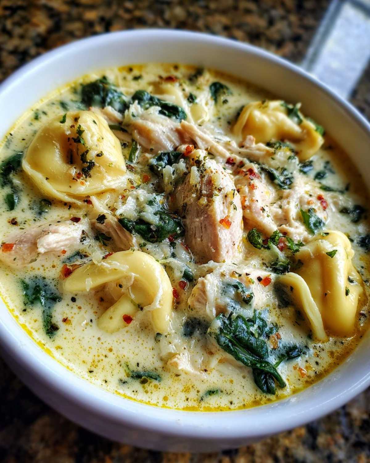 A close-up shot of a bowl of creamy chicken tortellini soup, featuring pasta, shredded chicken, and spinach, topped with herbs.