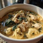 Close-up of a hearty bowl of creamy chicken tortellini soup featuring chunks of chicken, pasta, and wilted spinach.