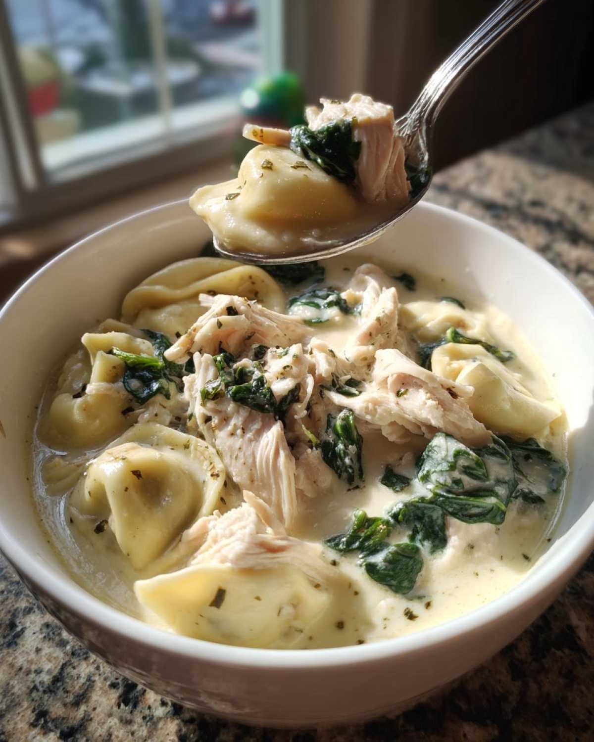 A spoonful lifting tortellini, chicken, and spinach from a bowl of creamy chicken tortellini soup.