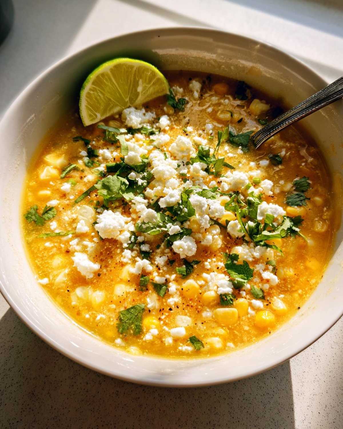 A close-up of a bowl of creamy corn soup topped with crumbled white cheese, cilantro, and a lime wedge.