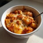 Close-up of a white bowl filled with cheesy, baked Creamy Roasted Garlic Tomato Gnocchi Bake, sprinkled with herbs.