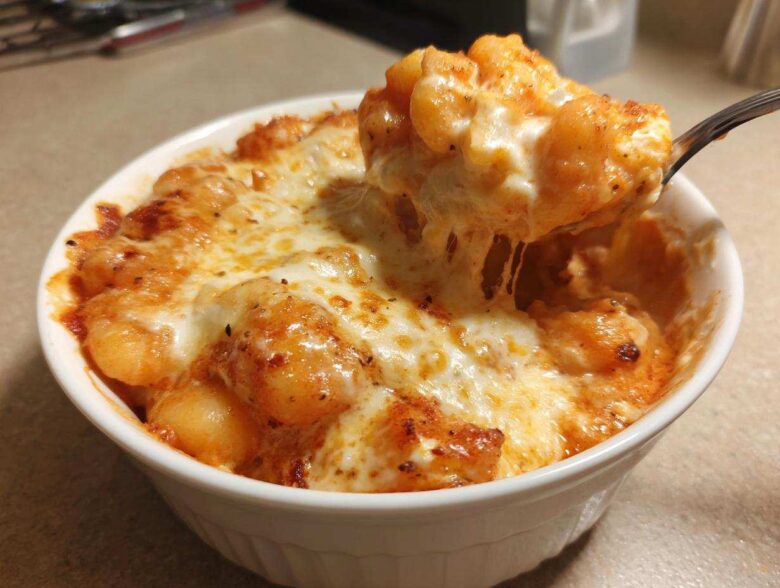 A spoonful of Creamy Roasted Garlic Tomato Gnocchi Bake being lifted from a white bowl, showing melted, stringy cheese.