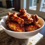 Close-up of crispy, glazed buffalo wings piled high in a white bowl on a granite countertop.