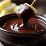 Close-up of an apple slice being dipped into a pot of smooth, melted chocolate fondue.
