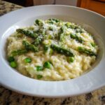 A close-up of a bowl of Easy Creamy Asparagus Risotto topped with bright green asparagus spears, peas, and grated Parmesan cheese.