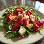 A close-up of an Easy Strawberry Avocado Salad featuring bright red strawberries, green avocado chunks, and crumbled white cheese over mixed greens.