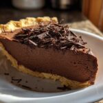 A close-up of a thick slice of french silk pie with a flaky crust, topped with dark chocolate shavings.