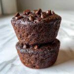 A close-up stack of two rich, dark chocolate muffins topped with melted chocolate chips.