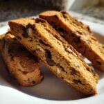Close-up of three slices of golden brown Gluten-Free Chocolate Chip Biscotti, studded with chocolate chips and almonds, resting on a white plate.