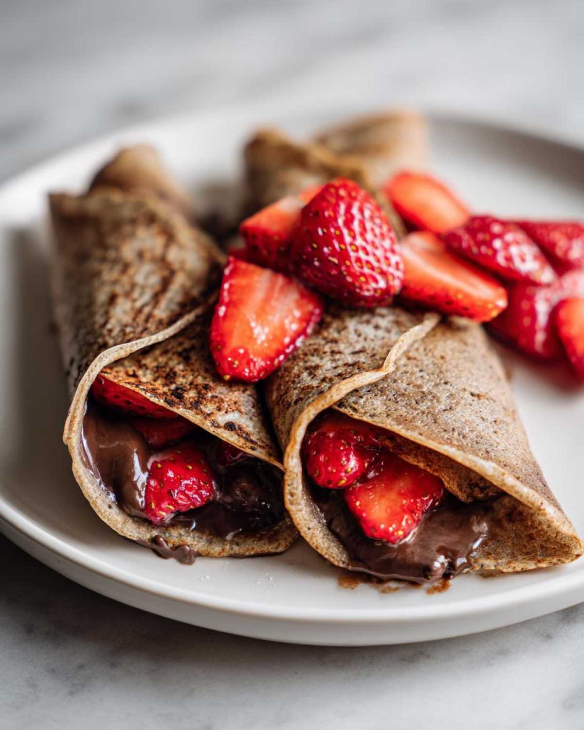 Two rolled Gluten-Free Chocolate Strawberry Crepes filled with chocolate spread and fresh strawberries on a white plate.