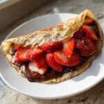 A folded Gluten-Free Chocolate Strawberry Crepes filled with rich chocolate spread and fresh sliced strawberries, dusted with powdered sugar.