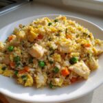 Close-up of a white plate piled high with Gluten-Free Lemon Pepper Fried Rice featuring chunks of chicken, scrambled egg, peas, and carrots.