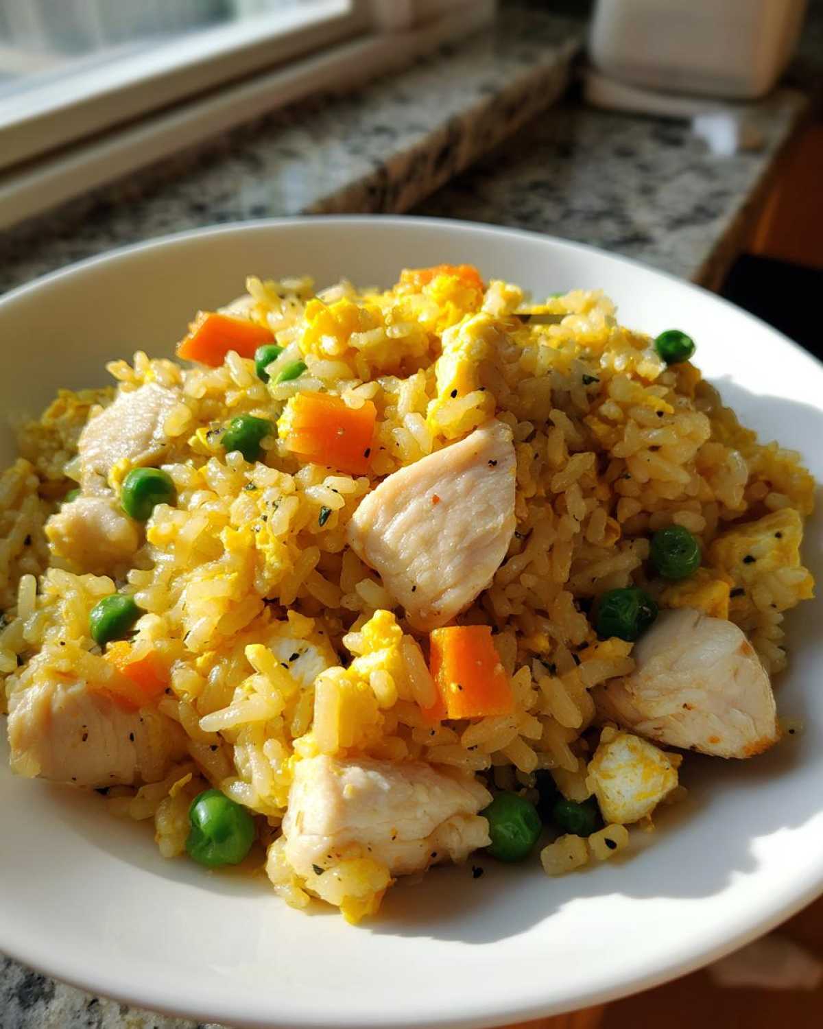 Close-up of a white bowl filled with Gluten-Free Lemon Pepper Fried Rice featuring chunks of chicken, scrambled egg, peas, and carrots.