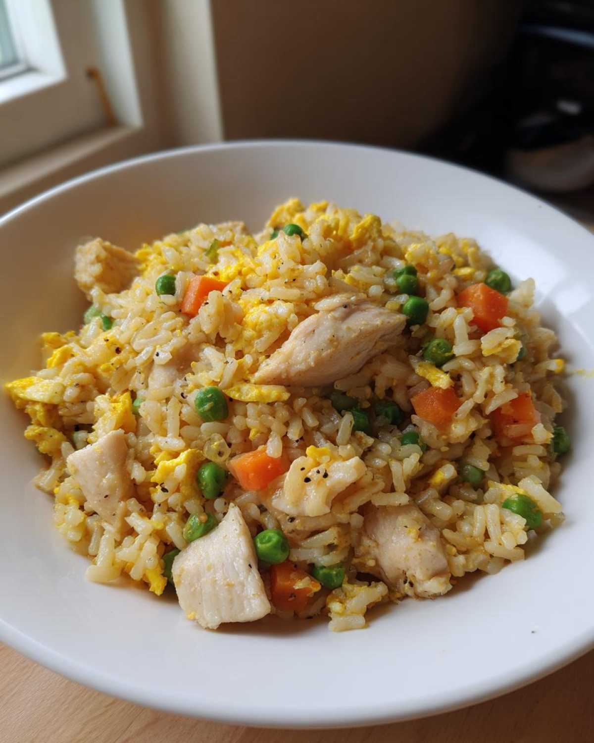 Close-up of a white bowl filled with Gluten-Free Lemon Pepper Fried Rice featuring chicken pieces, scrambled egg, peas, and carrots.