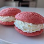 Two pink Gluten-Free Strawberry Lemon Whoopie Pies filled with thick white cream, resting on a white plate.