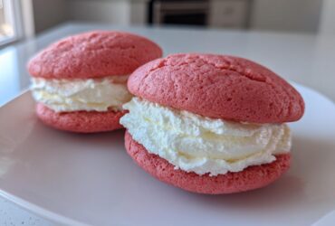 Two pink Gluten-Free Strawberry Lemon Whoopie Pies filled with thick white cream, resting on a white plate.