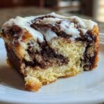 A close-up of a single, gooey slice of cinnamon roll cake topped with white icing.