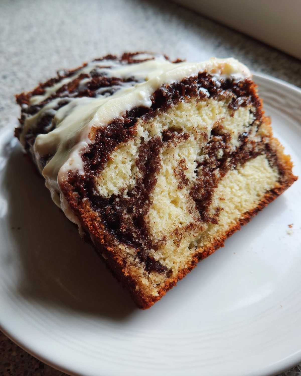 Close-up of a slice of marble-swirled cinnamon roll cake topped with thick, white cream cheese icing.