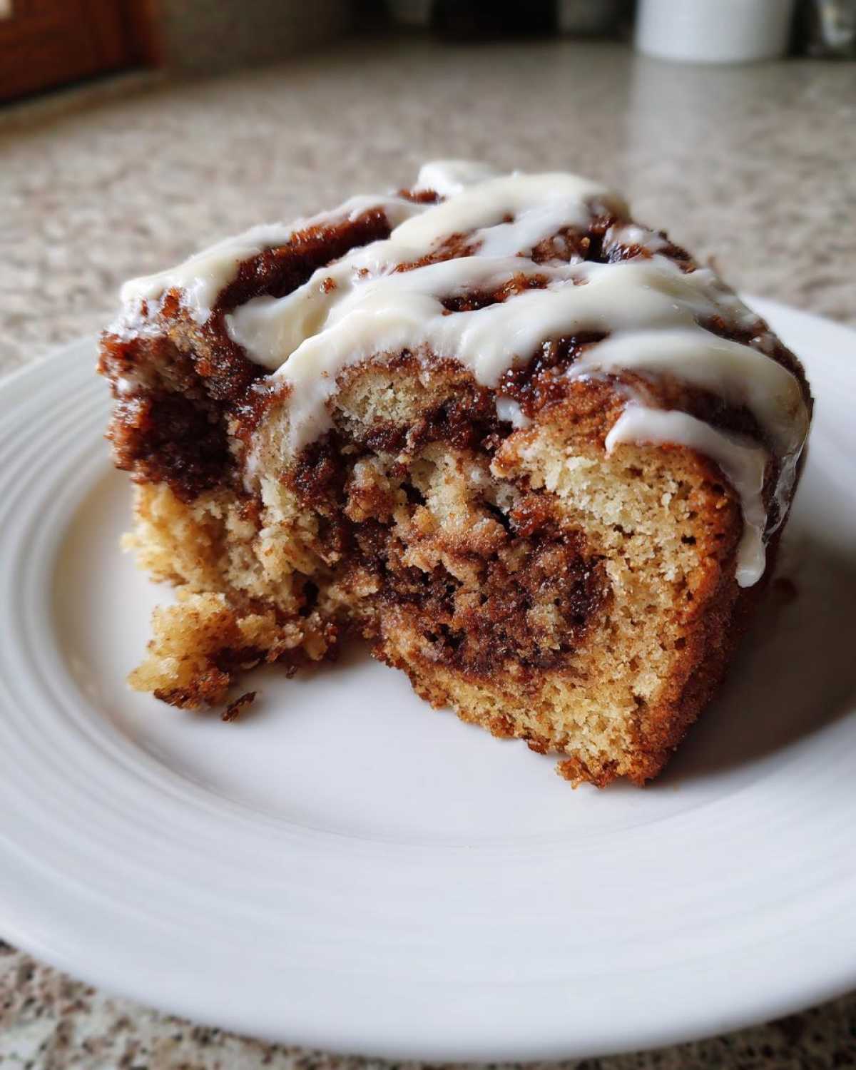 A close-up of a moist slice of cinnamon roll cake with a thick cinnamon swirl and drizzled with white icing.