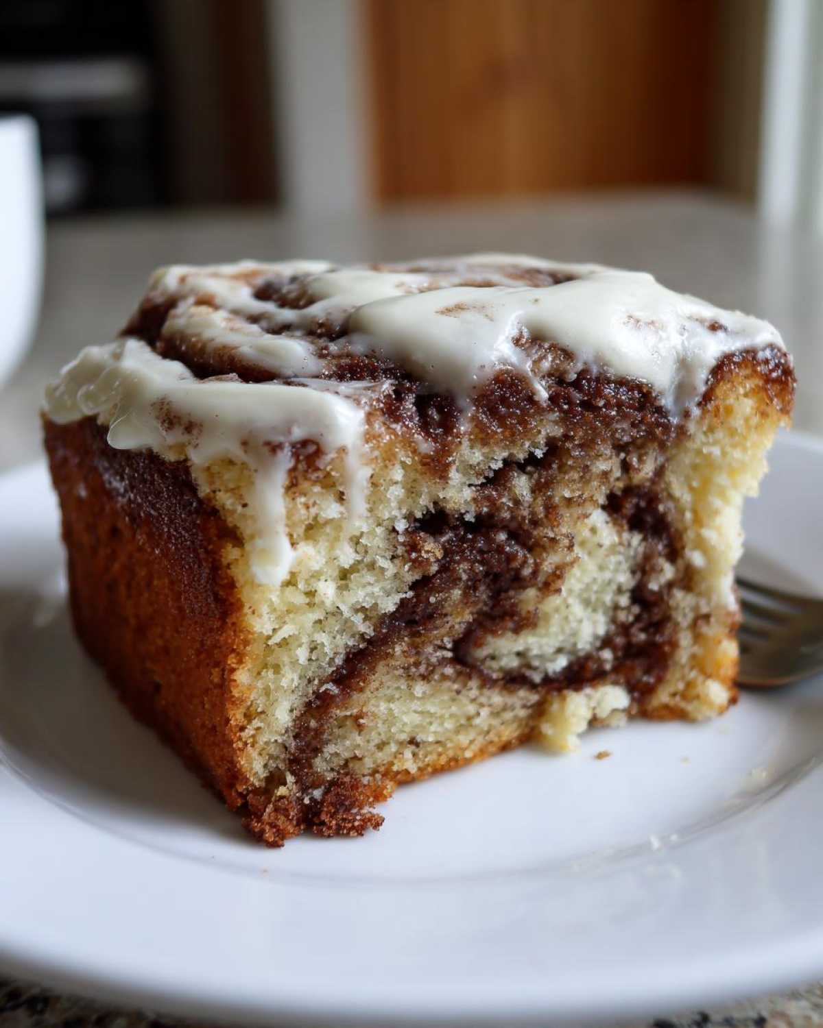 A moist slice of cinnamon roll cake featuring a swirl of cinnamon filling and topped with thick white icing.
