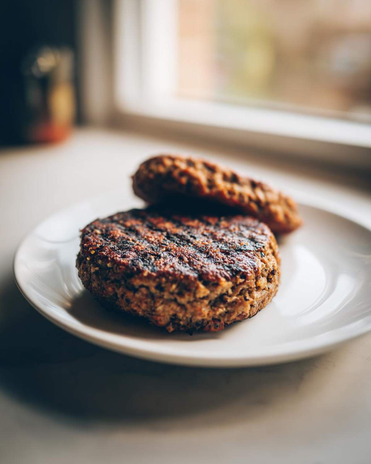 Two grilled black bean burger patties, one resting on top of the other, on a white plate near a window.