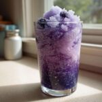 A tall glass filled with a vibrant purple Iced Blueberry Lemonade Slush, topped with shaved ice and whole blueberries.