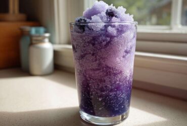 A tall glass filled with a vibrant purple Iced Blueberry Lemonade Slush, topped with shaved ice and whole blueberries.