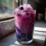 A tall glass filled with a vibrant, frosty Iced Blueberry Lemonade Slush, topped with crushed ice and whole blueberries.