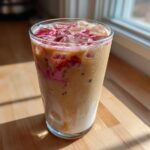 A tall glass of Iced Raspberry Vanilla Chai with ice cubes and bright pink raspberry syrup swirls, sitting on a wooden counter near a window.