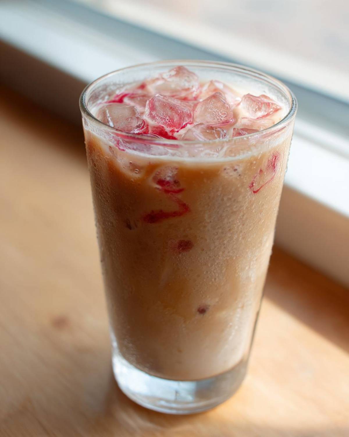 A tall glass filled with Iced Raspberry Vanilla Chai, showing condensation and ice cubes topped with raspberry syrup swirls.