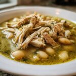 Close-up of a white bowl filled with Instant Pot Chicken Pozole Verde, featuring shredded chicken and white hominy in a green broth.