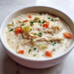 A close-up of a white bowl filled with Instant Pot Creamy Chicken and Rice Soup, showing shredded chicken, rice, carrots, celery, and parsley.
