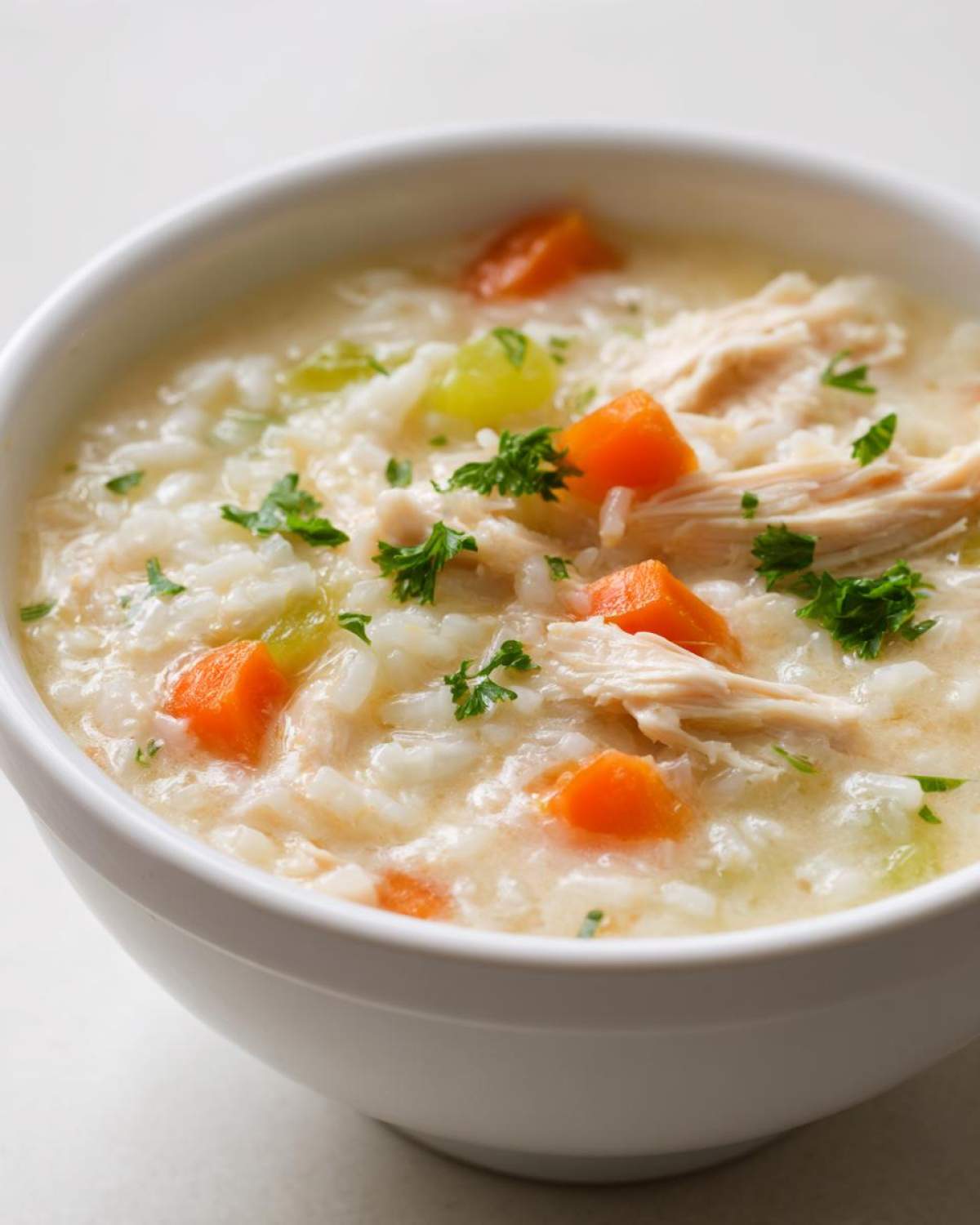 A close-up of a white bowl filled with Instant Pot Creamy Chicken and Rice Soup, showing shredded chicken, rice, carrots, and parsley garnish.