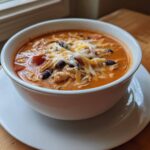 A white bowl filled with rich, orange Instant Pot Creamy Chicken Enchilada Soup, topped with shredded cheese, black beans, and chicken.