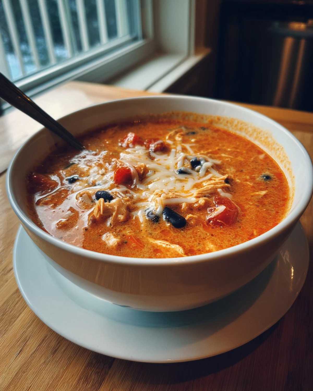 A close-up of a white bowl filled with Instant Pot Creamy Chicken Enchilada Soup, topped with shredded cheese, chicken, and black beans.