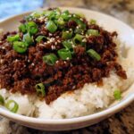 A close-up of a white bowl filled with white rice topped generously with saucy Instant Pot Korean Beef and garnished with green onions and sesame seeds.