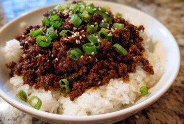 A close-up of a white bowl filled with white rice topped generously with saucy Instant Pot Korean Beef and garnished with green onions and sesame seeds.