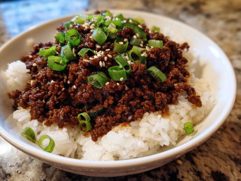 A close-up of a white bowl filled with white rice topped generously with saucy Instant Pot Korean Beef and garnished with green onions and sesame seeds.