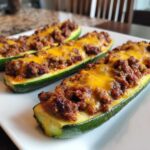 Three Keto Cheesy Taco Stuffed Zucchini Boats filled with seasoned ground meat and melted cheddar cheese on a white plate.