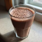 A glass filled with a thick, frothy Keto Chocolate Strawberry Smoothie sitting near a sunny window.