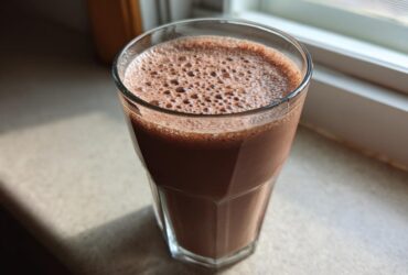 A glass filled with a thick, frothy Keto Chocolate Strawberry Smoothie sitting near a sunny window.