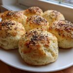 Six freshly baked Keto Everything Bagel Cloud Bread buns topped with seeds, arranged on a white plate near a window.