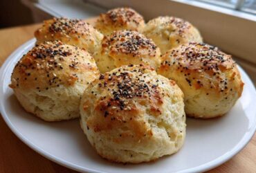 Six freshly baked Keto Everything Bagel Cloud Bread buns topped with seeds, arranged on a white plate near a window.